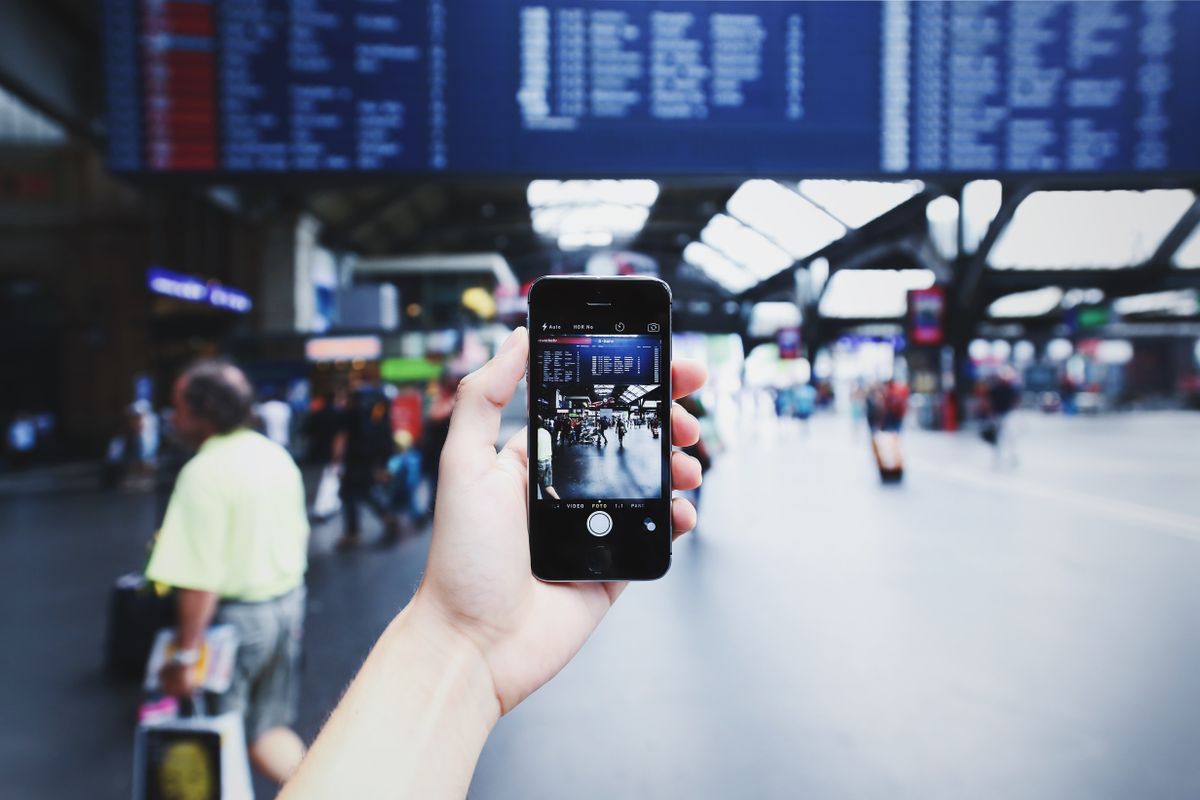 Mao segurando smartphone diante do painel de horarios em estacao, representando conectividade em viagem