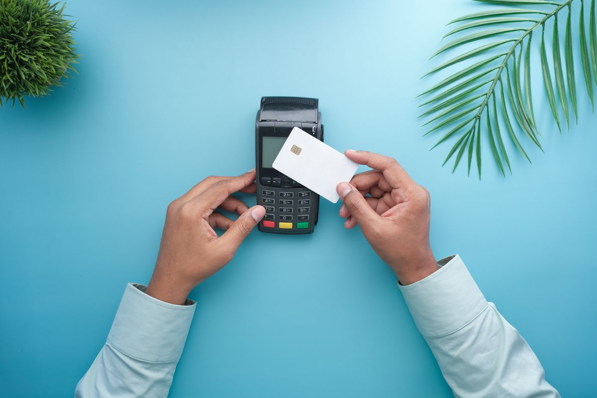 Mãos aproximando um cartão de uma maquininha de pagamento sobre fundo azul, representando o split payment nas viagens corporativas