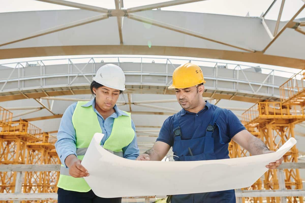 Engenheira e engenheiro de capacete analisando uma planta em canteiro de obras, representando viagens corporativas na construção civil